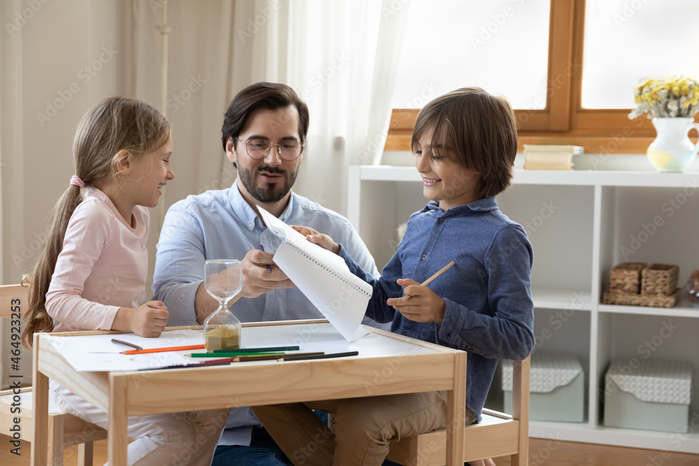 Smiling millennial father teaching small children son daughter writing letters or drawing pictures in paper album, sitting at table with sand clock, enjoying spending creative time together at home. Smiling millennial father teaching small children son daughter writing letters or drawing pictures in paper album, sitting at table with sand clock, enjoying spending creative time together at home.