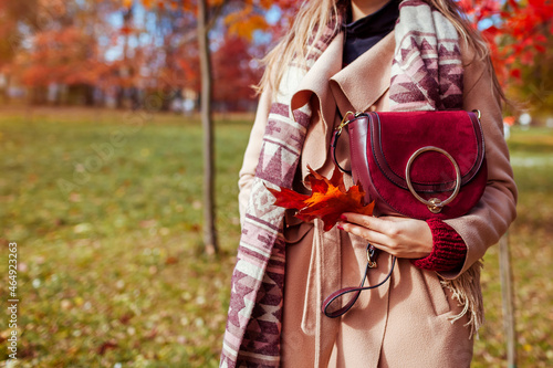 Close up of stylish woman holding burgundy purse wearing coat in autumn park. Fall female clothes, accessories. Fashion