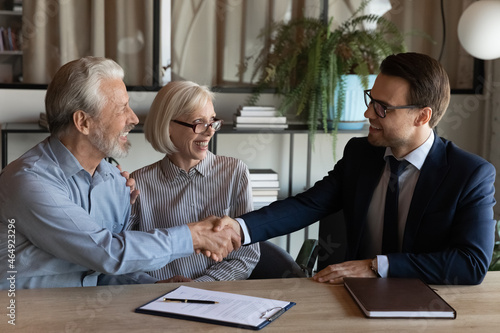 Shaking hands. Smiling young man real estate broker handshaking with older family couple after buying selling property. Spouses retirees thank bank agent for help after signing loan mortgage agreement
