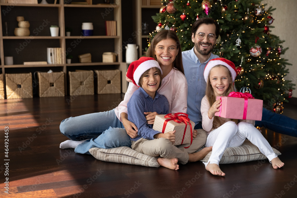 Portrait of happy loving young couple parents holding on laps laughing small children siblings sitting on pillows on floor near decorated Christmas tree with wrapped gift boxes, celebration concept.