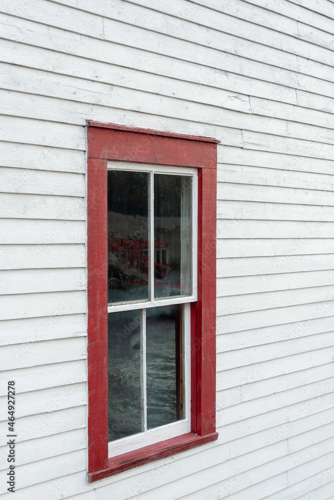 Fototapeta premium The exterior of an old white wooden shed wall with a red framed closed glass double hung window. In the window is a reflection of the sky and you can see green rope stacked inside the building.