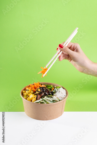 A woman's hand with chopsticks over a salad poke bowl and tuna, carrot, cucumber and pineapple with a green background.