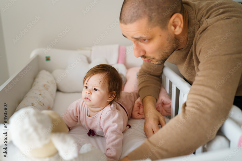 Baby girl lying in cradle bed on belly with her father taking care of her caucasian cute infant baby play in a crib in bright room at home growing up parenthood concept