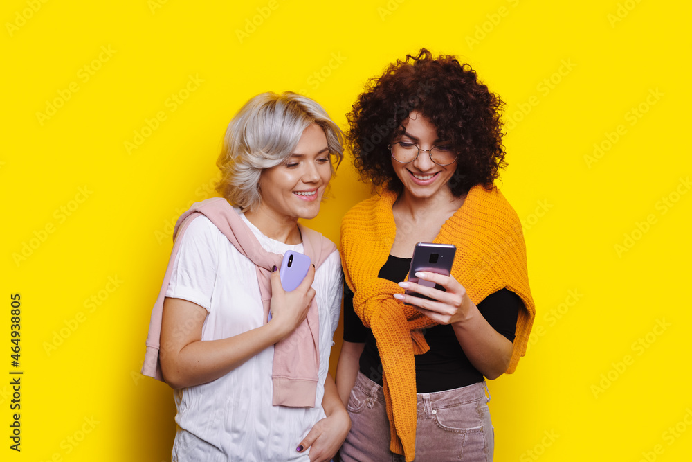 Woman with curly hair looks at something on the phone while woman with blond and short hair peeks. Yellow background