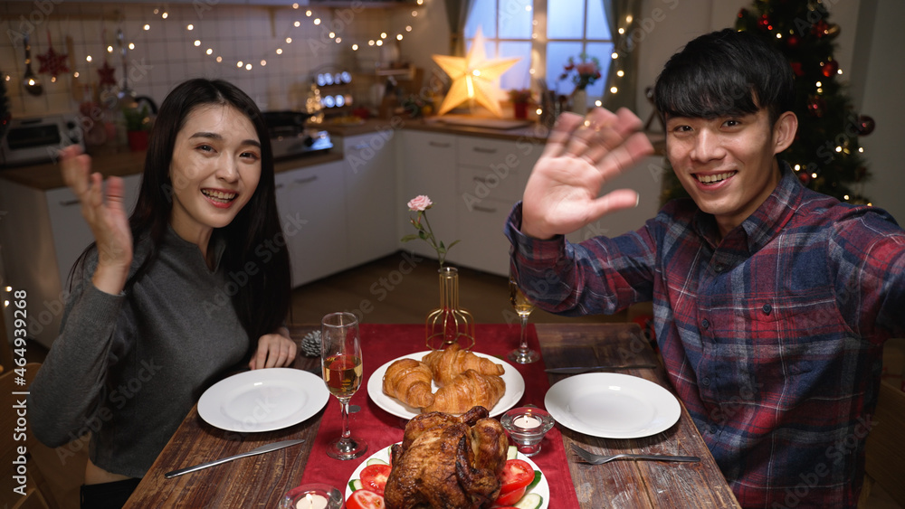 young asian couple enjoy dinner at home on xmas eve. asian man and woman face camera having video call greetings to parents online. male and female celebrate Merry Christmas of holiday near the Tree 