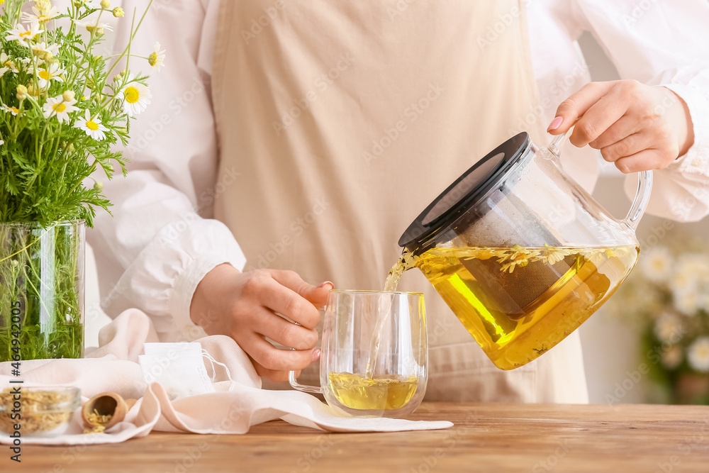 Woman pouring tasty chamomile tea from teapot into cup on table Woman pouring tasty chamomile tea from teapot into cup on table