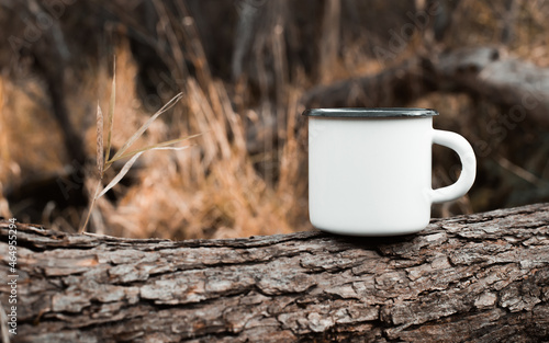 Camping metal white mug mockup standing on tree in forest outdoors, copy space for text. Enamelled empty cup for branding or logo