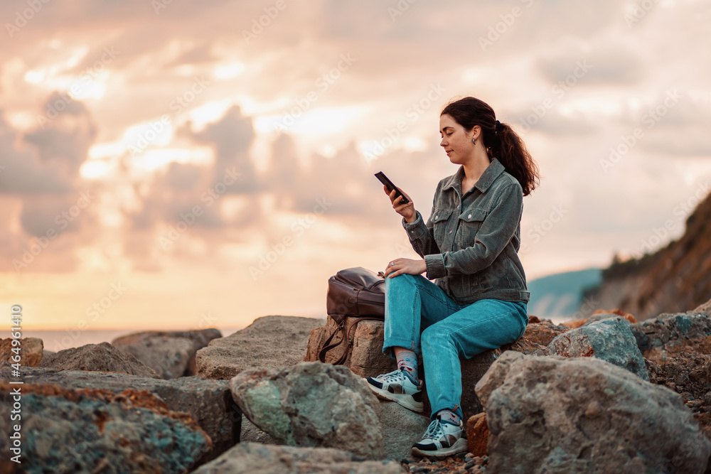 A young woman is sitting on rocky rocks and using a phone. Sunset in the background. Communication concept