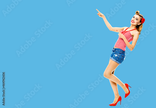 Full body photo of happy excited smiling woman pointing at something. Blond girl in pin up style, showing copy space area for text or imaginary. Retro fashion and vintage. Blue color background.