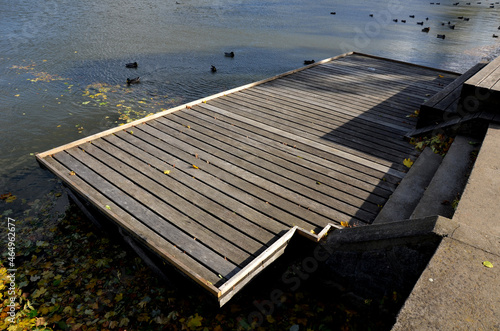 Pier for park visitors. two steps lead to it. planks terrace over water. Pond with a dock for boats. floor plan of a rectangle. the terrace has no railings.