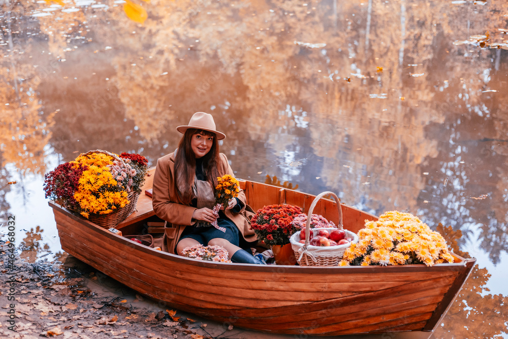 young beautiful plus size model in autumn brown warm coat and hat sitting in boat on pond decorate with chrysanthemums and apples in autumn park, concept of season leaf fall, romance and unusual date