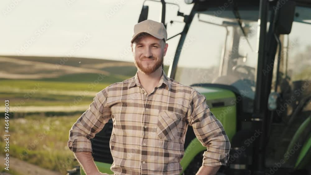 A young bearded farmer is putting on a cap and keeping his hands on his belt. He is smiling and looking at the camera. He is standing in the middle of a field near a tractor. The sun is shining. 4K A young bearded farmer is putting on a cap and keeping his hands on his belt. He is smiling and looking at the camera. He is standing in the middle of a field near a tractor. The sun is shining. 4K