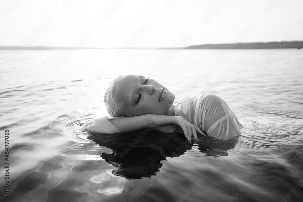 Fototapeta premium Beautiful blonde woman short hair lies in water on sea shore, pond. Art portrait of a relaxing woman in the water on shore