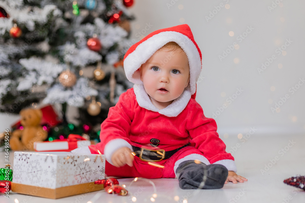 baby in a Santa costume is sitting next to the Christmas tree and playing with gift boxes. child opens a Christmas gift. winter new year's concept. space for text. High quality photo
