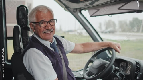 Medium portrait of cheerful senior bus driver in uniform sitting at driver seat with hands on steering wheel smiling to camera photo