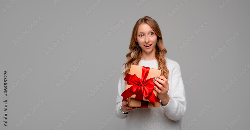 Fototapeta premium Beautiful young woman holding in hands unboxed present and looking at camera with surprised expression, standing over gray background. Holidays, celebration and lifestyle concept. Mock up copy space