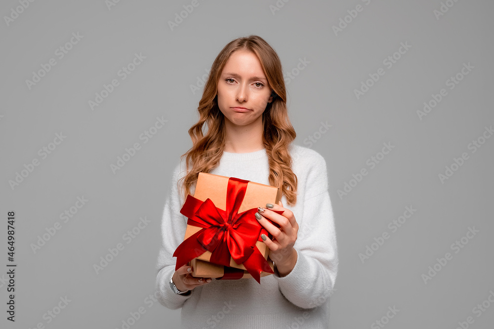 Young woman in white sweater holding gift box looking at camera with displeased upset expression, standing over gray background Young woman in white sweater holding gift box looking at camera with displeased upset expression, standing over gray background