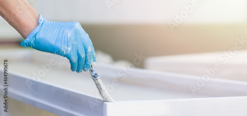 Male hands in blue gloves hold a paintbrush and paint a wooden board with white paint outdoors. The man is engaged in construction work and self-repair. Horizontal format banner. Close-up. Copy space