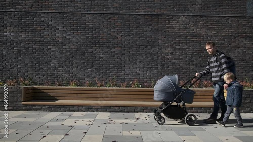 Father and his little son walking with newborn baby stroller on the city near brick wall. Man in maternity leave with pram.
