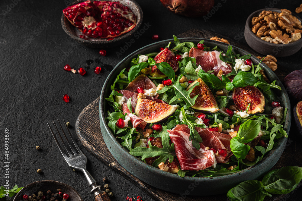 Autumn salad with figs, prosciutto, baby rocket, goat cheese, nuts and pomegranate seeds with fork on black background