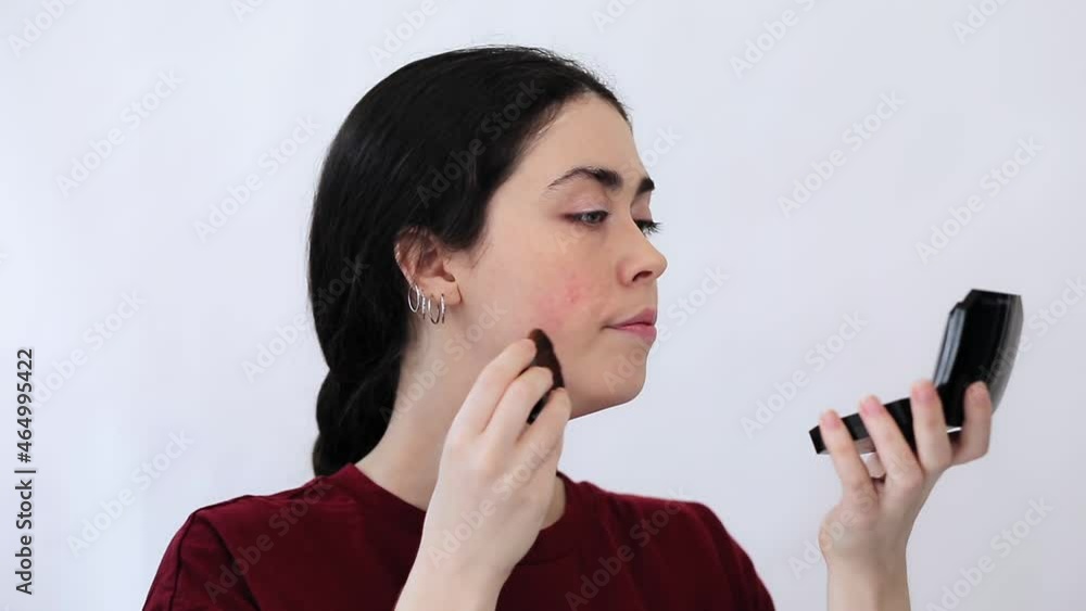 Portrait of a young woman applying powder with a brush, masking redness on her cheeks with cosmetics. White background. The concept of rosacea and couperose. Portrait of a young woman applying powder with a brush, masking redness on her cheeks with cosmetics. White background. The concept of rosacea and couperose.