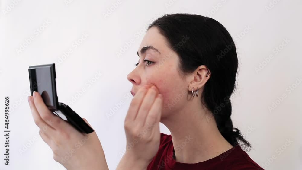 Close-up portrait of a young woman applying powder with a brush, masking redness on her cheeks with cosmetics. White background. The concept of rosacea and couperose. Close-up portrait of a young woman applying powder with a brush, masking redness on her cheeks with cosmetics. White background. The concept of rosacea and couperose.