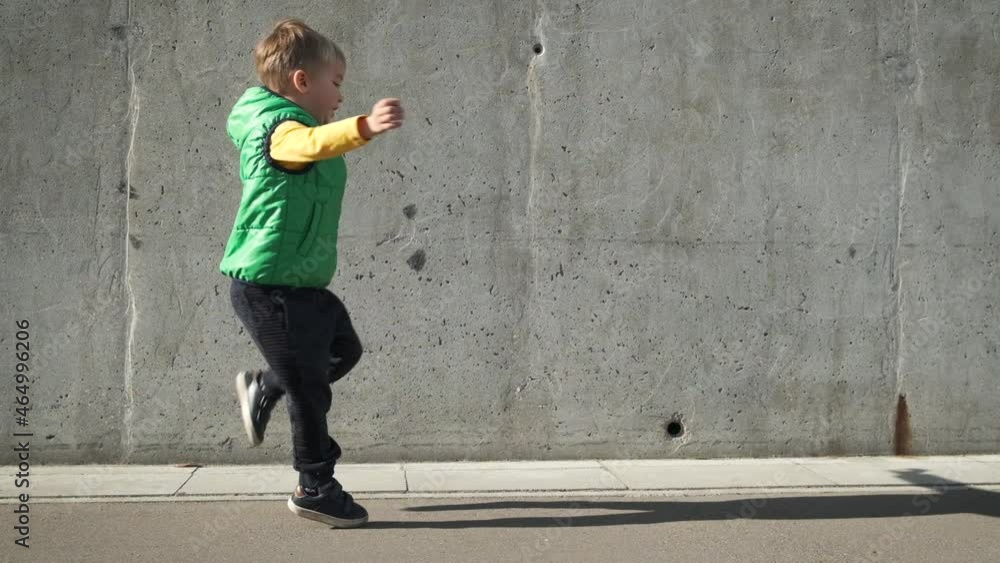 Cool modern preschool child boy in yellow and green clothes dancing near grey concrete wall in the city in sunny day. Funny ecstatic kid jumping and having fun in slow motion. Cool modern preschool child boy in yellow and green clothes dancing near grey concrete wall in the city in sunny day. Funny ecstatic kid jumping and having fun in slow motion.