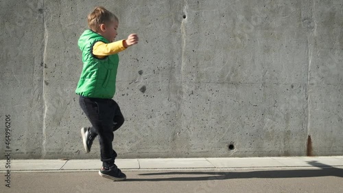 Cool modern preschool child boy in yellow and green clothes dancing near grey concrete wall in the city in sunny day. Funny ecstatic kid jumping and having fun in slow motion.