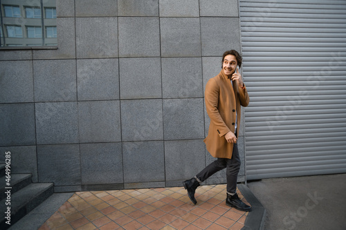 Cheerful man in brown coat talking on smartphone and walking on urban street  © Dmytro Hai