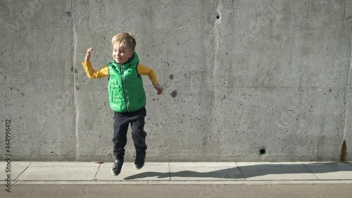 Cool trendy modern preschool child boy in yellow and green clothes dancing near grey concrete wall in the city in sunny day. Funny ecstatic kid jumping and having fun in slow motion.