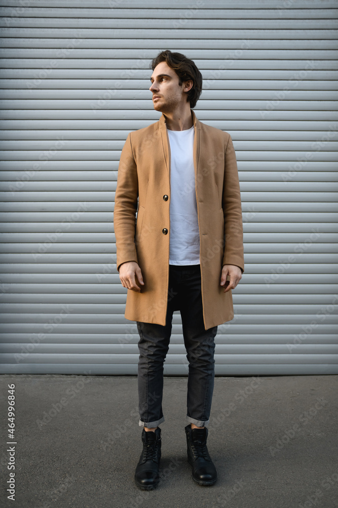 Full length of stylish man in coat and boots standing near roller shutter outdoors Full length of stylish man in coat and boots standing near roller shutter outdoors