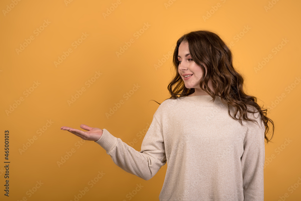 A young and attractive Caucasian brunette girl with wavy hair holding a empty space in her palm on an orange studio background.