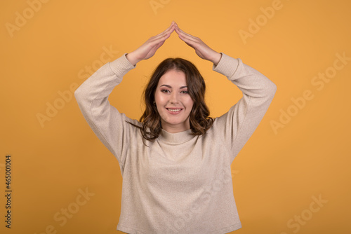 A young and attractive Caucasian brunette girl with wavy hair showing the roof of the house with her hands on an orange studio background.