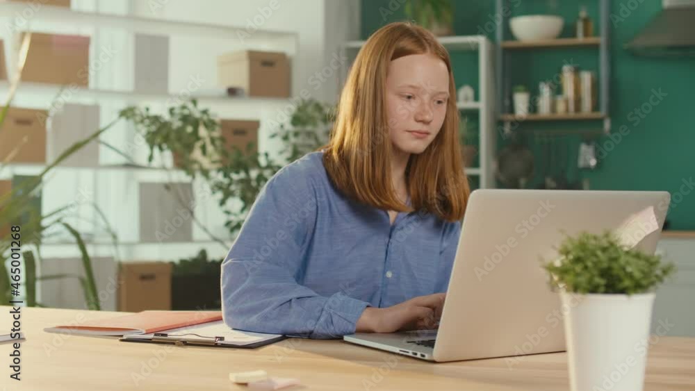 Red-haired Teenage Girl Studying at Home Online, Watching a Video Tutorial, Doing Homework while Sitting at a Table With a Laptop. Education and Virtual Distance Learning. Virus, Quarantine. Red-haired Teenage Girl Studying at Home Online, Watching a Video Tutorial, Doing Homework while Sitting at a Table With a Laptop. Education and Virtual Distance Learning. Virus, Quarantine.
