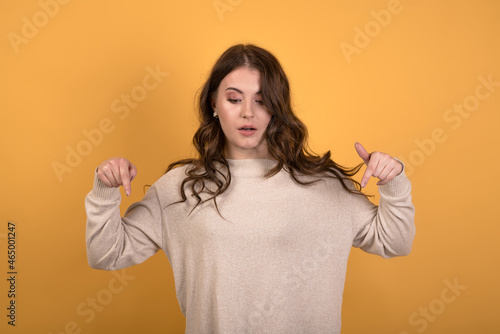 A young and attractive Caucasian brunette girl with wavy hair in a casual jumper pointing her fingers down while showing an advertisement on an orange studio background.