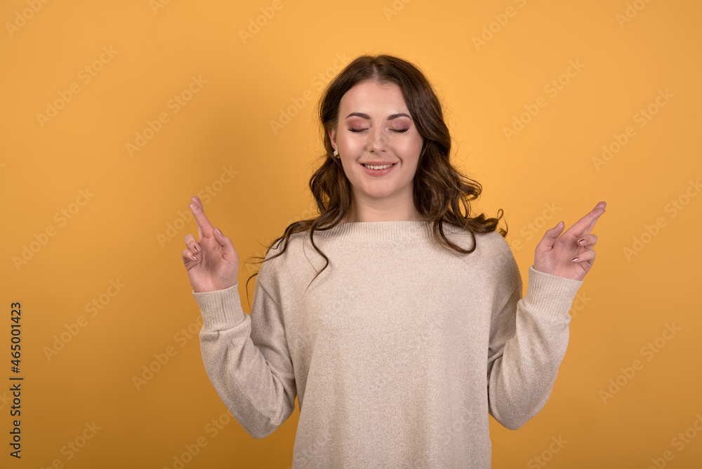 Portrait of a happy and attractive Caucasian brunette girl with wavy hair in a casual jumper with crossed fingers on an orange studio background. Luck and trust. Portrait of a happy and attractive Caucasian brunette girl with wavy hair in a casual jumper with crossed fingers on an orange studio background. Luck and trust.