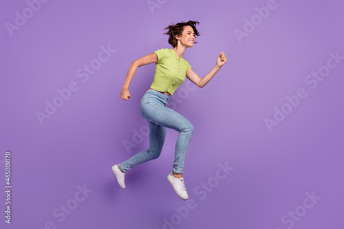 Photo of positive funky young woman dressed green t-shirt jumping high running fast looking empty space isolated purple color background