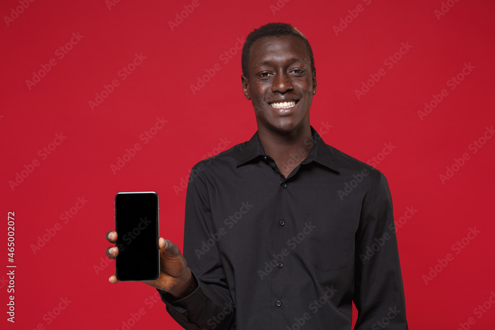 Smiling young african american man guy in classic black shirt posing isolated on red wall background in studio. People lifestyle concept. Mock up copy space. Hold mobile phone with blank empty screen. Smiling young african american man guy in classic black shirt posing isolated on red wall background in studio. People lifestyle concept. Mock up copy space. Hold mobile phone with blank empty screen.