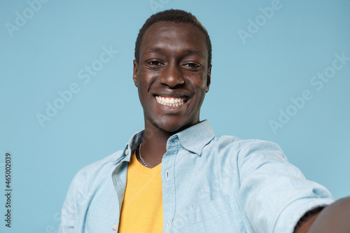 Close up of smiling young african american man guy in casual shirt, yellow t-shirt posing isolated on blue background. People lifestyle concept. Mock up copy space. Doing selfie shot on mobile phone.