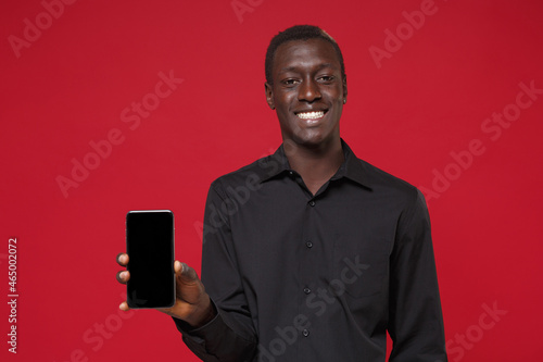 Smiling young african american man guy in classic black shirt posing isolated on red wall background in studio. People lifestyle concept. Mock up copy space. Hold mobile phone with blank empty screen. © ViDi Studio