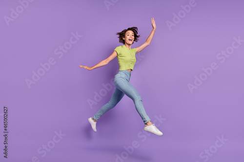 Photo of charming excited young woman dressed green t-shirt jumping walking waving arm empty space isolated purple color background