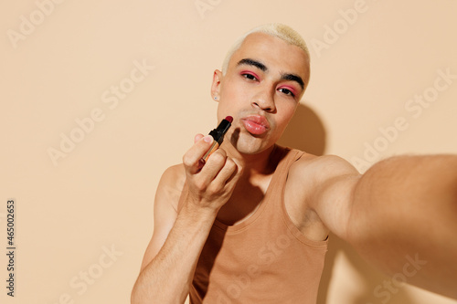 Close up sexy latin gay man with make up in beige tank shirt do selfie shot pov mobile phone painting lips with lipstick isolated on plain light ocher background studio People lgbt lifestyle concept.