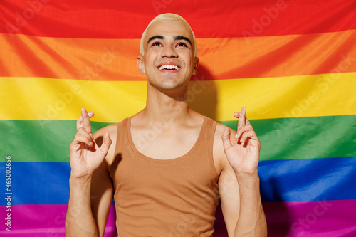 Side view young queer smiling blond latin gay man with make up in beige tank shirt waiting for special moment  keeping fingers crossed on rainbow flag background studio People lgbt lifestyle concept.