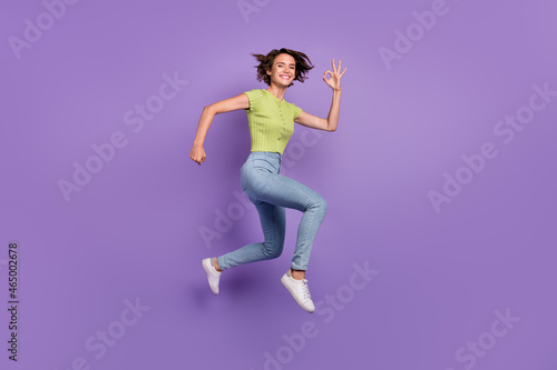 Photo of impressed sweet young lady wear green t-shirt showing fingers okey jumping high isolated violet color background