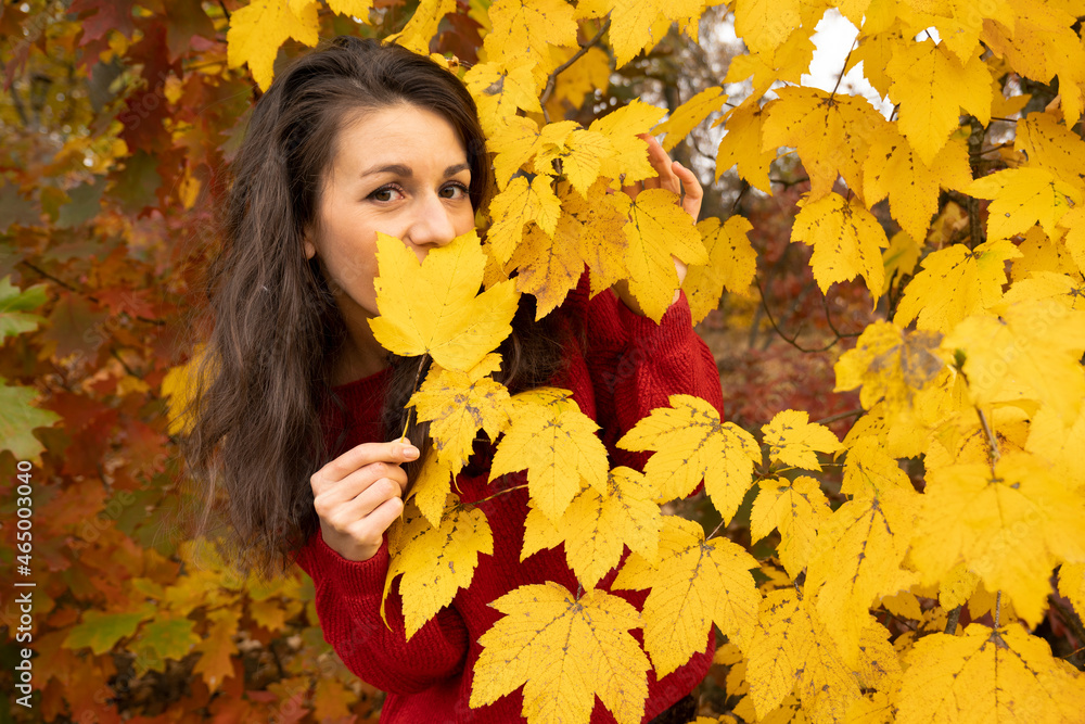 Young woman peeking out from behind yellow foliage in autumn
