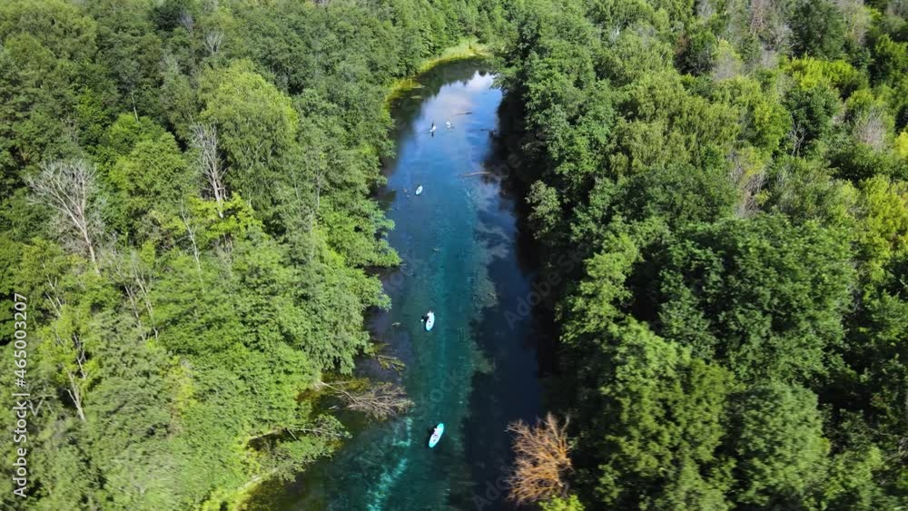 Summer hot day. A group of tourists is paddling down river on paddle boards. The banks of the winding rivulet are overgrown with tall trees with lush green foliage. Calm water of deep turquoise color. Summer hot day. A group of tourists is paddling down river on paddle boards. The banks of the winding rivulet are overgrown with tall trees with lush green foliage. Calm water of deep turquoise color.