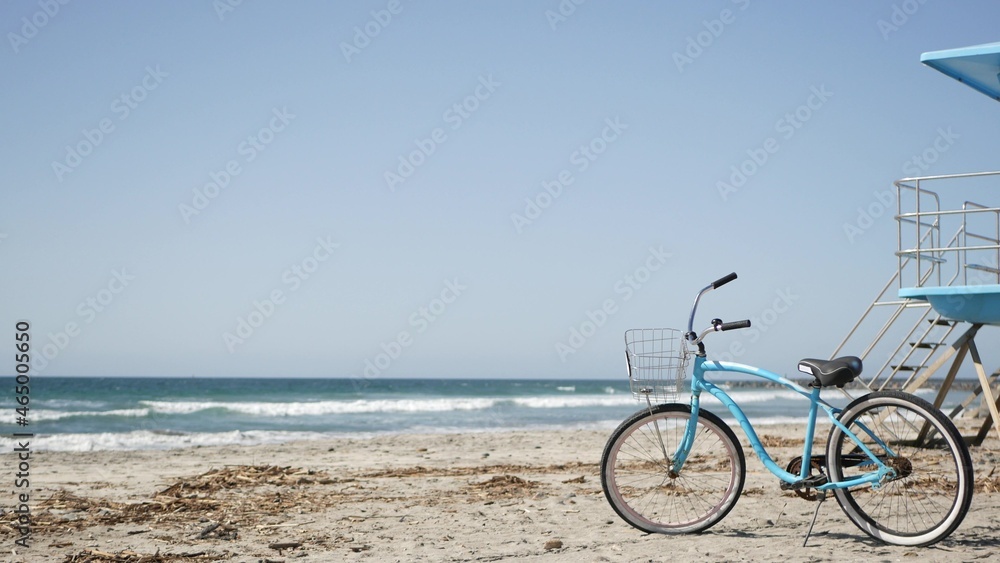 Blue bicycle, cruiser bike by ocean beach, pacific coast, Oceanside California USA. Summertime vacations, sea shore. Vintage cycle on sand near lifeguard tower or watchtower hut. Sky and water waves.