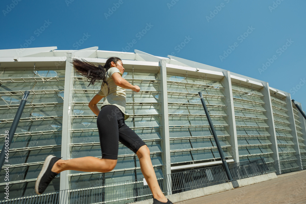 Slim young Asian woman in comfortable tracksuit runs along road past modern stadium building under clear sky on sunny day