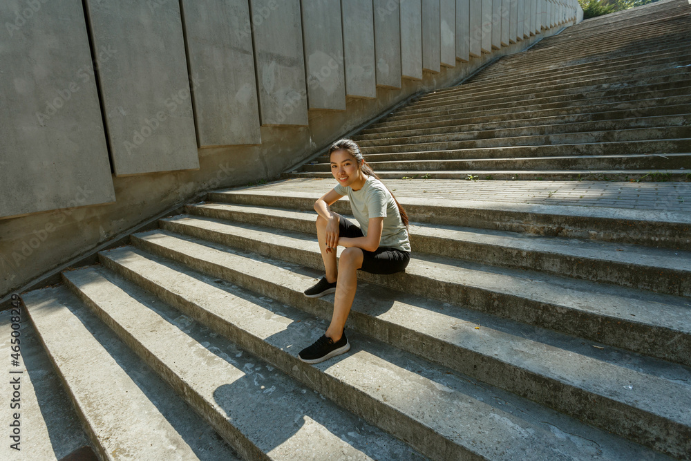 Attractive Asian woman in tracksuit and sneakers sits resting on empty grey steps casting shadow after outdoor training in sunny city Attractive Asian woman in tracksuit and sneakers sits resting on empty grey steps casting shadow after outdoor training in sunny city