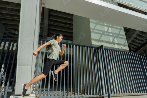 Graceful sportive Asian woman in stylish tracksuit jumping upward moment at training near modern multi-storey parking fence on sunny day © sergiymolchenko